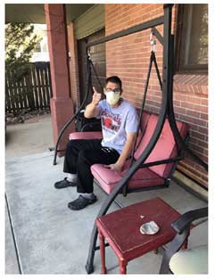 A teenage boy sitting on a swinging settee on his porch, waving.