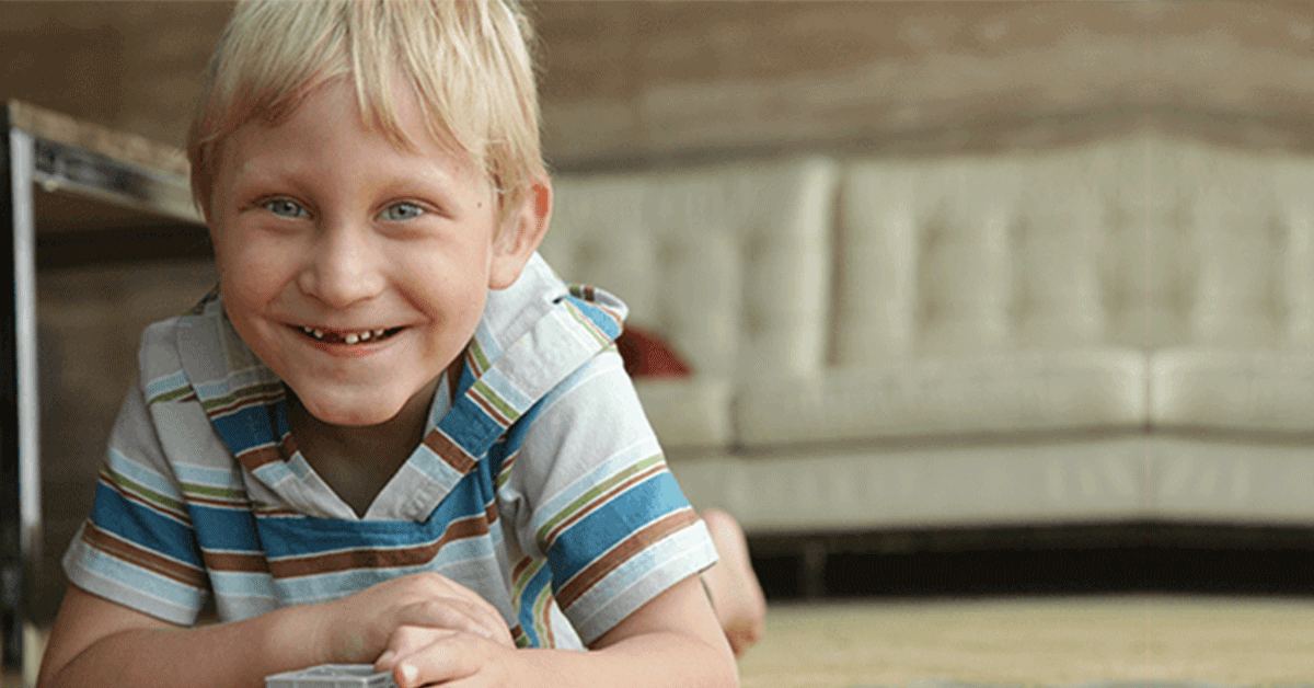 A young blonde boy on the floor smiling at the camera.