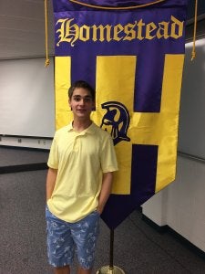 Teenage boy in short sleeves and shorts standing in front of a banner reading Homestead