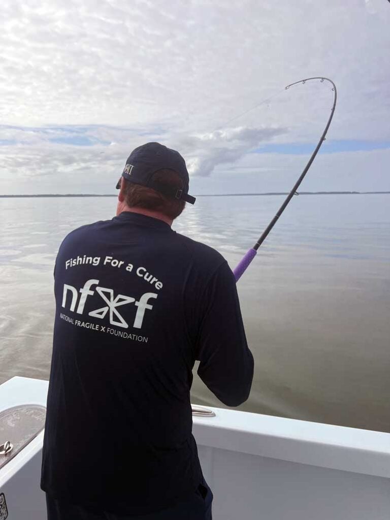 Casting a line into the ocean, a man facing away is wearing a shirt that says Fishing for a Cure.