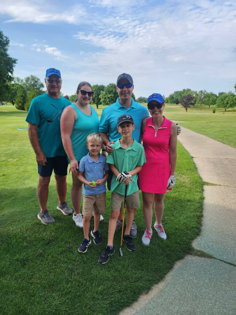 Four adults and two young boys on a golf course on a sunny day.