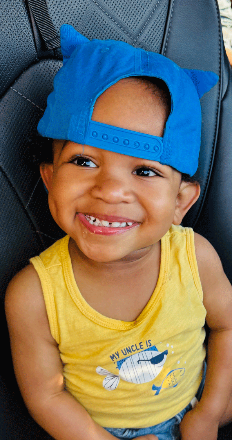 Camden Adjmul wearing a backwards baseball hat, sitting in his car seat, and showing the most beautiful smile ever.