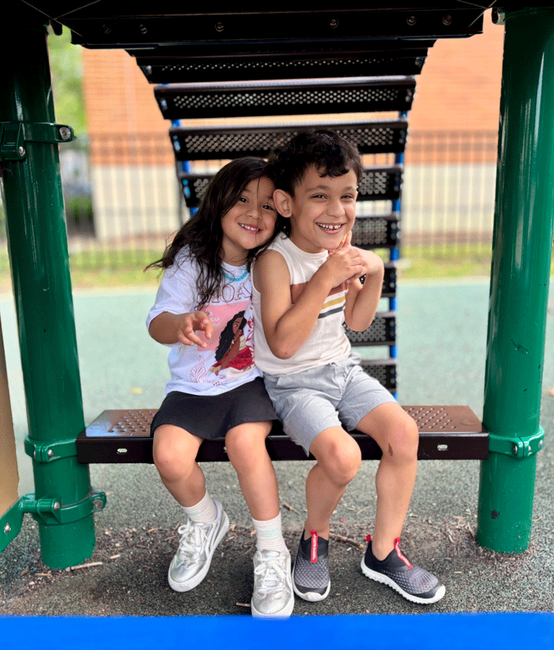 Jojo and Pia at a playground, sitting together closely and smiling big.