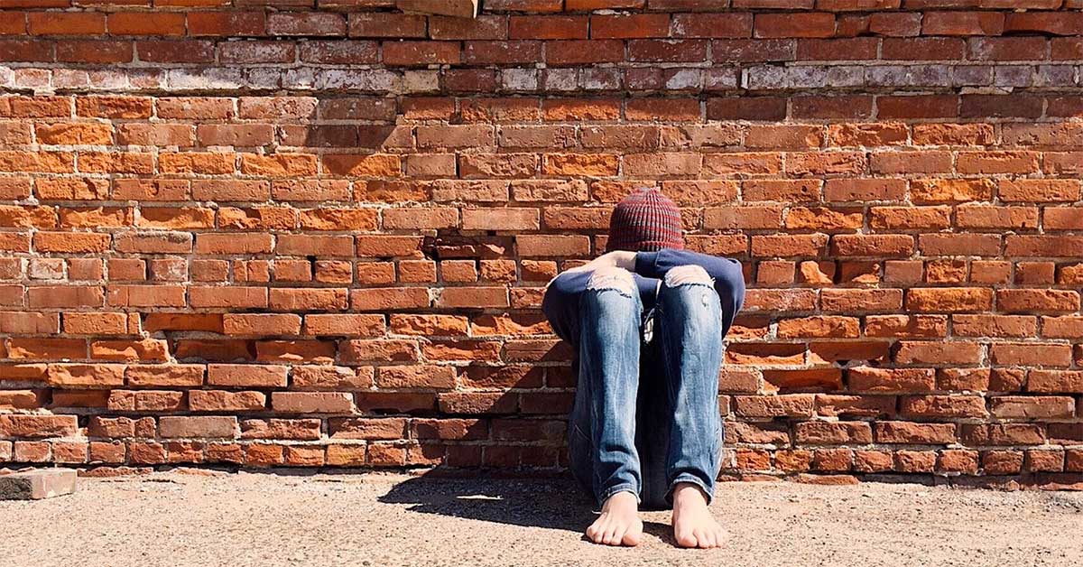 A teen sitting on a sidewalk with his head down.