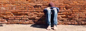 A teen sitting on a sidewalk, his knees bent and he's leaning against a brick wall. His head is down and his arms are crossed.