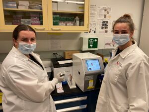 Two females in white lab coats with the blood spot analysis machine.