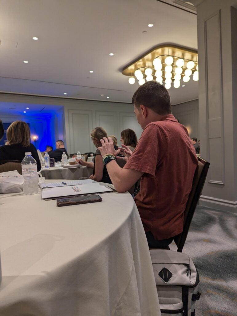 A man with a red shirt sitting at a table listening to a speaker