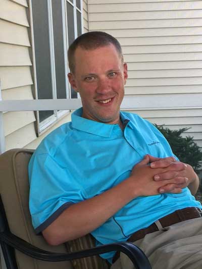 A young man who has Fragile X syndrome, Denny Haugen, wearing a turquoise shirt and a winning smile sitting on his front porch.