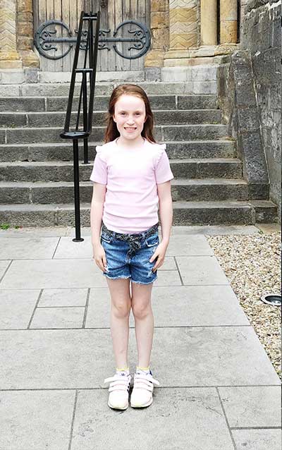 A young girl, Alannah, standing with very good posture on a walkway, and wearing a pink t-shirt, denim shorts, and pink shoes.