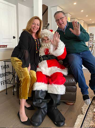 Andrew Pritchard, an adult with Fragile X syndrome, dressed as Santa and waving with his mom and dad sitting on each arm of the chair he's sitting in.