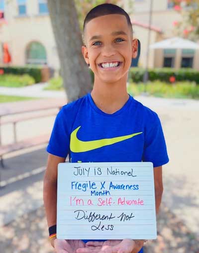 Audric Newsome, a young boy with Fragile X syndrome, smiling big and holding a sign reading "July 18 National Fragile X Awareness Month; I'm a Self-Advocate; Different - Not Less."