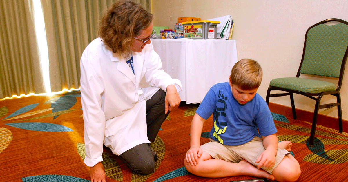 A clinician in a white lab coat kneeling on the floor next to a young boy wearing shorts sitting on the floor. She is engaging with the boy.