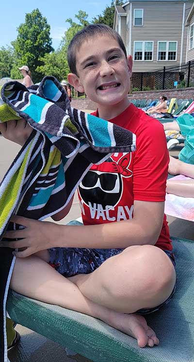 A young boy who has Fragile X syndrome, Ayden, outdoors on a beach among other beachgoers wearing swim clothes and a delightful smile.