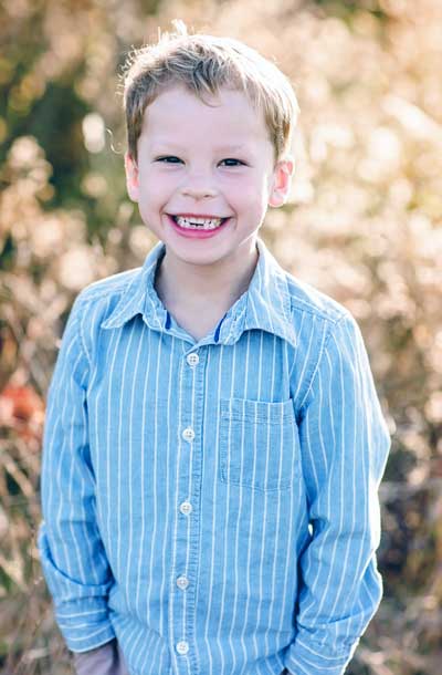 Benjamin Smith, a young boy with Fragile X syndrome, outdoors wearing a striped blue dress shirt and a big happy smile.