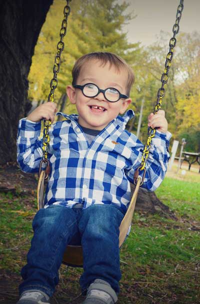 Bramel Maggard, a young boy with Fragile X syndrome, on a tree swing wearing glasses, plaid shirt, jeans, and super happy smile.
