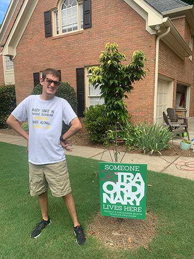 A young man Bryan Muma on his front lawn next to a sign that reads SOMEONE XTRAORDINARY LIVES HERE.