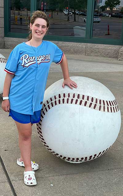 Caragan Raska wearing a Rangers jersey and standing next to a huge 3-foot baseball.