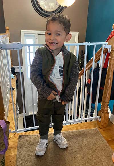 A young boy, Carter, standing on a stairway landing indoors and wearing tennis shoes, dark pants, a white t-shirt, gray jacket, and a big smile.