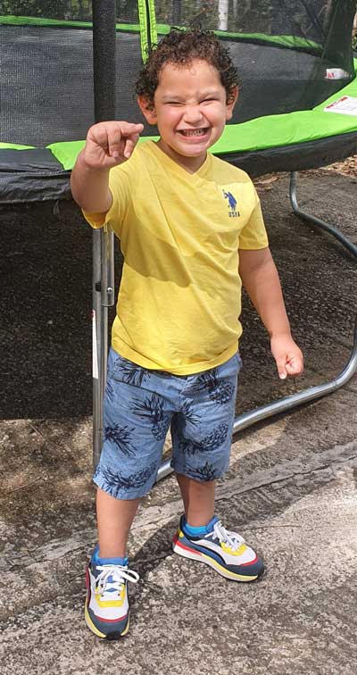 A young boy who has Fragile X syndrome, Cesar, wearing a yellow t-shirt, blue shorts, and a big smile, standing in front of a trampoline.