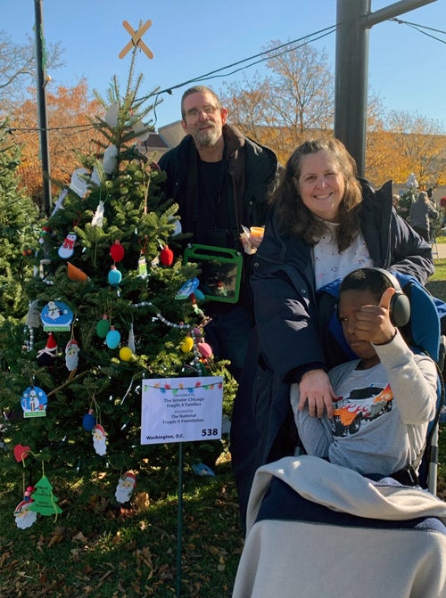 A man, woman, and young boy in a wheelchair giving a thumbs up, next to their decorated Christmas tree.