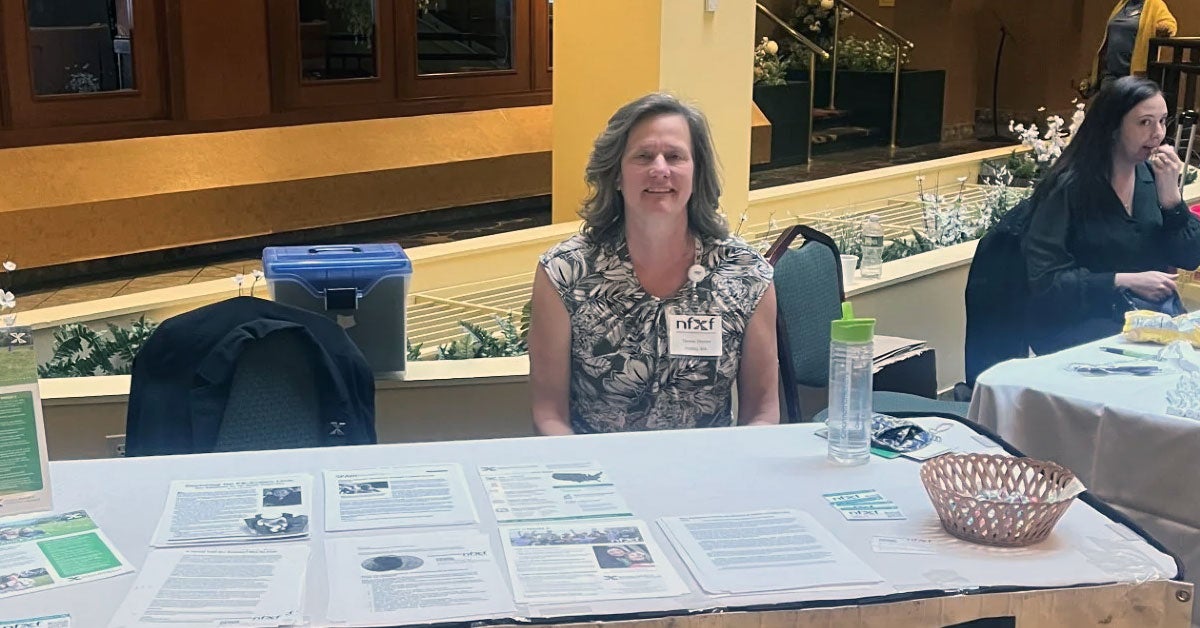 Denise Devin sitting behind a Fragile X information display table at the Autism Connections annual conference.