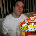 A middle-aged brunette male sitting at a table wearing a white t-shirt with a cake and gifts on the table before him