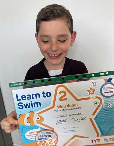 A young boy who has Fragile X syndrome, Gael Centellas, smiling and holding a huge "Learn to Swim" award for "Well done!"