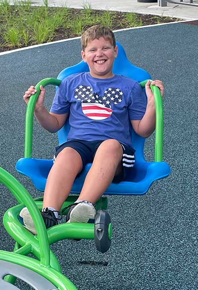 A young boy Grayson is wearing a t-shirt and shorts and sitting on a playground apparatus.