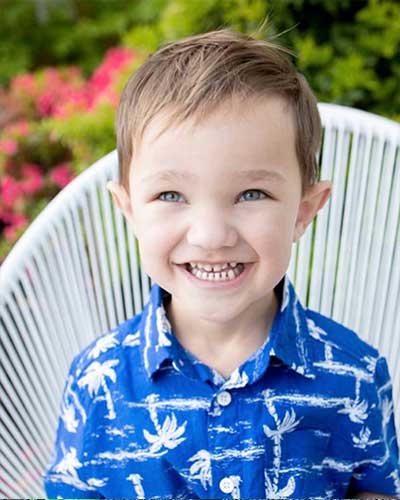 A young boy Gus Hansen sitting on a white chair and wearing a tropical blue and white shirt, and a huge smile.