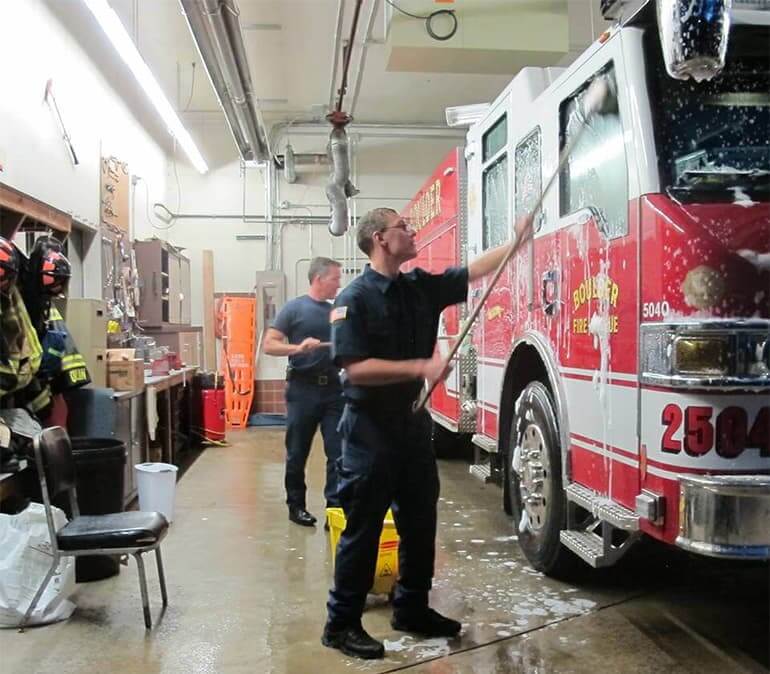 2 men washing a fire truck with long brushes and soap and water