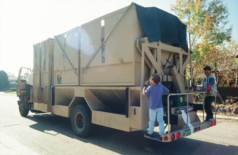 A boy and a man riding on the back of a garbage truck.