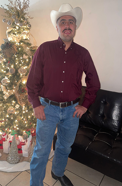 Jose in front of a Christmas tree wearing jeans, boots, and a cowboy hat.