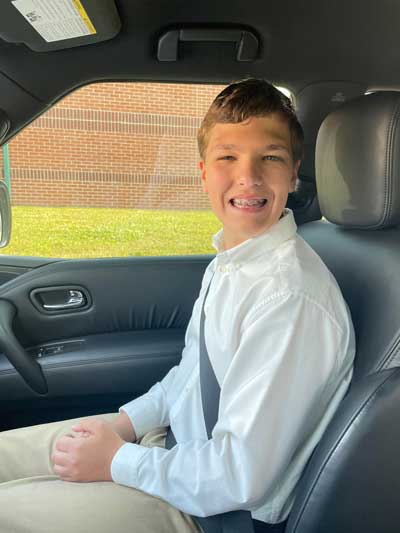 Kaiden, a young man with Fragile X syndrome, sitting in the passenger seat of a car and wearing a white dress shirt, khaki pants, and a charming smile.