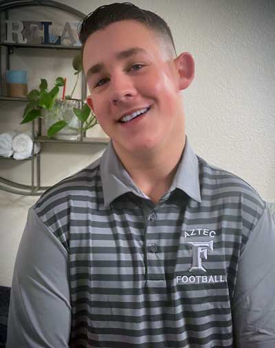 Kolton Sanders, a young man with Fragile X syndrome, wearing an Aztec Football collared shirt and a winning smile.