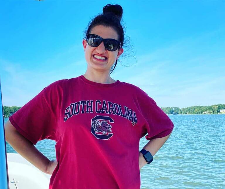 Mallory wearing sunglasses and posing by a lake, smiling big