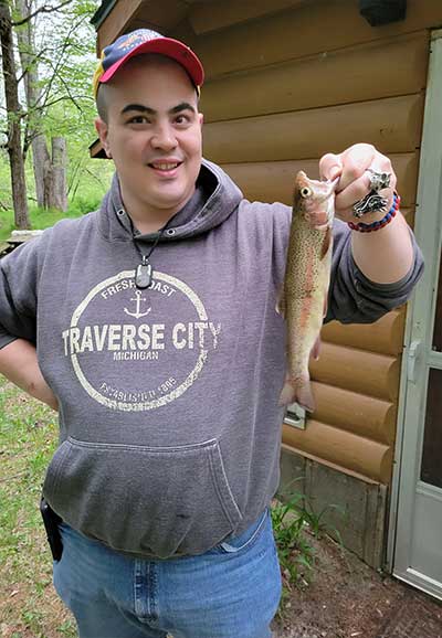 Nathan Hesling outdoors wearing jeans, a sweatshirt, and baseball cap, holding a fish he just caught.