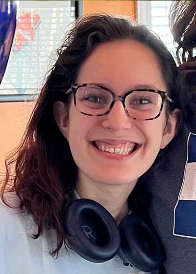 A young woman Nicole Brooke Kuperman with dark hair, tortoise shell glasses, and headphones around her neck.