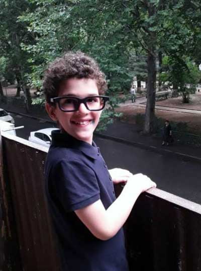 Nicoloz, a young boy with Fragile X syndrome, wearing glasses and a dark blue polo shirt, standing on a balcony with his hands on the railing.