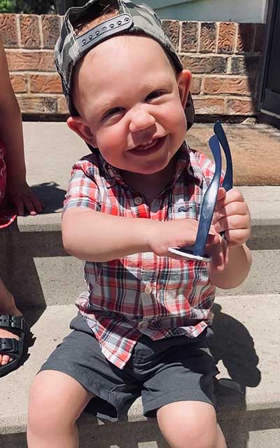 Toddler Paxton sitting on a step and wearing shorts, a plaid shirt, and a baseball cap on backwards; holding a pair of sunglasses and and huge smile.