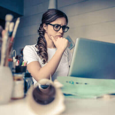Young adult brunette woman with a long braid and black-rimmed glasses scowling at a laptop computer on her desk and surrounded by pen and pencils in cups
