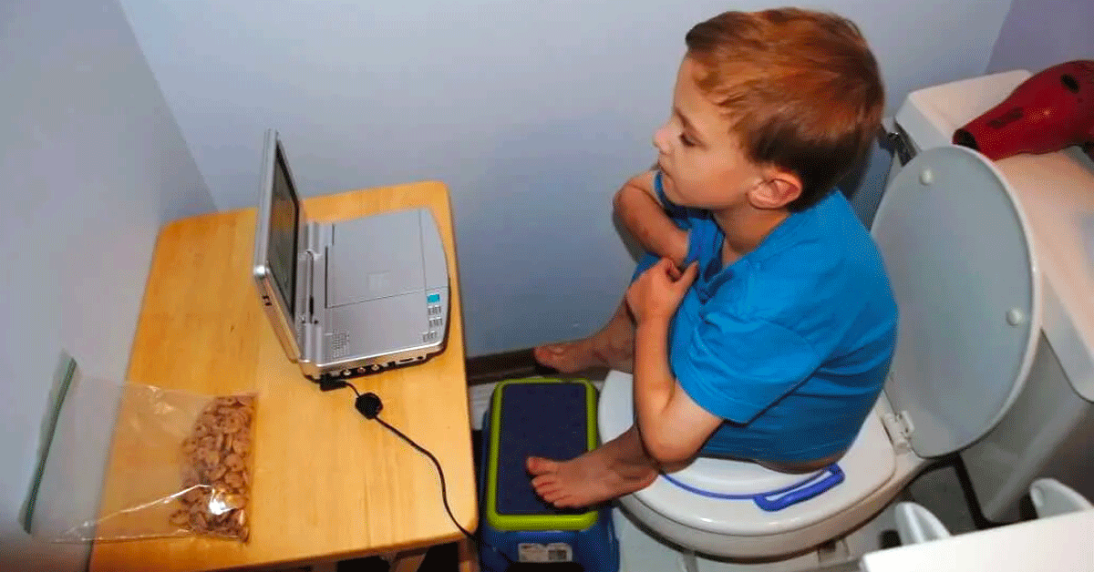 Young boy going through toilet training, sitting on a toilet with a laptop computer and snacks.
