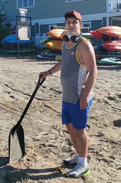 Tyler Krantz, a young man with Fragile X syndrome, wearing a tank top and shorts with headphones around his neck and standing with a shovel at a marina.